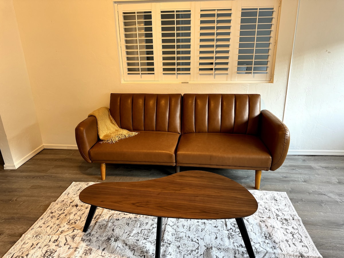 A comfortable couch with a tan leather finish is featured, complemented by a textured throw draped over one arm. A uniquely shaped wooden coffee table sits in front, resting on a patterned area rug. Light from a nearby window enhances the cozy setting.