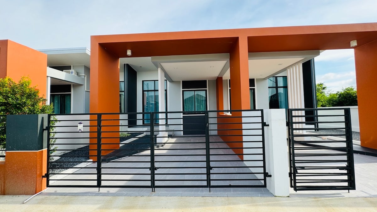 The entrance features a modern design with a prominent orange facade and sleek black gates. Large windows allow natural light to enter the interior space. The paved pathway leads to the main door, framed by neatly maintained greenery and a landscaped area.