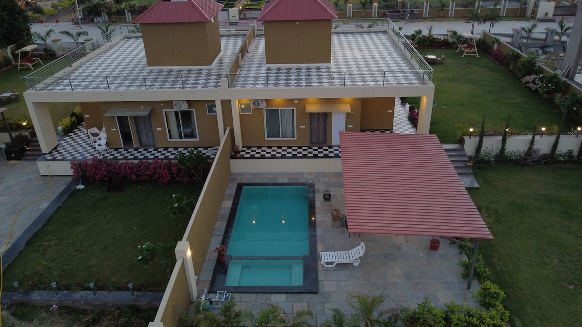 An aerial view of a well-maintained villa showcases a private pool in the center, surrounded by a landscaped garden. The property features a spacious terrace with distinct rooftops and sleek outdoor furniture. Lush greenery and blooming flowers enhance the inviting outdoor space.