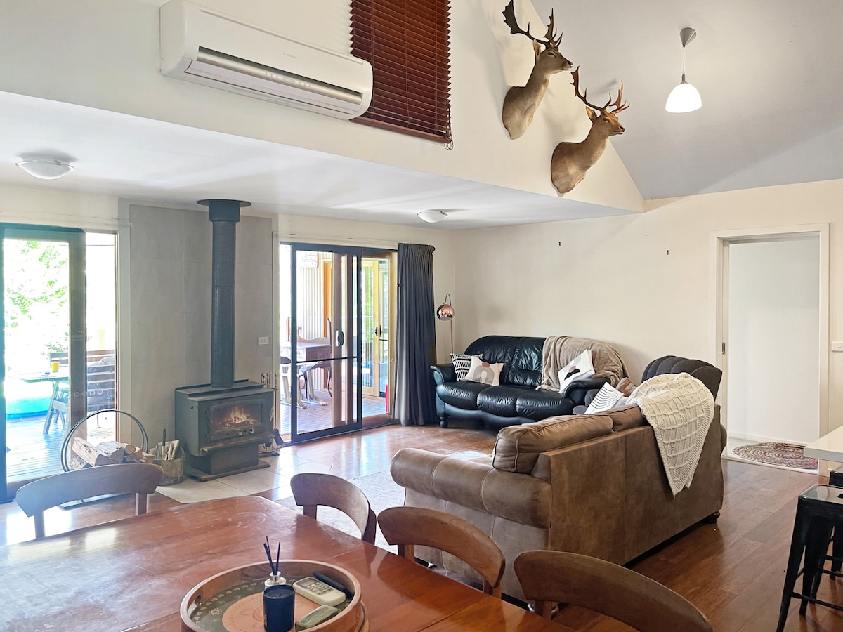 A spacious lounge area features two comfortable sofas, one brown with a throw blanket, and a black one, arranged around a wooden coffee table. A fireplace provides warmth, while doors lead to the outdoor deck, allowing natural light to fill the room.