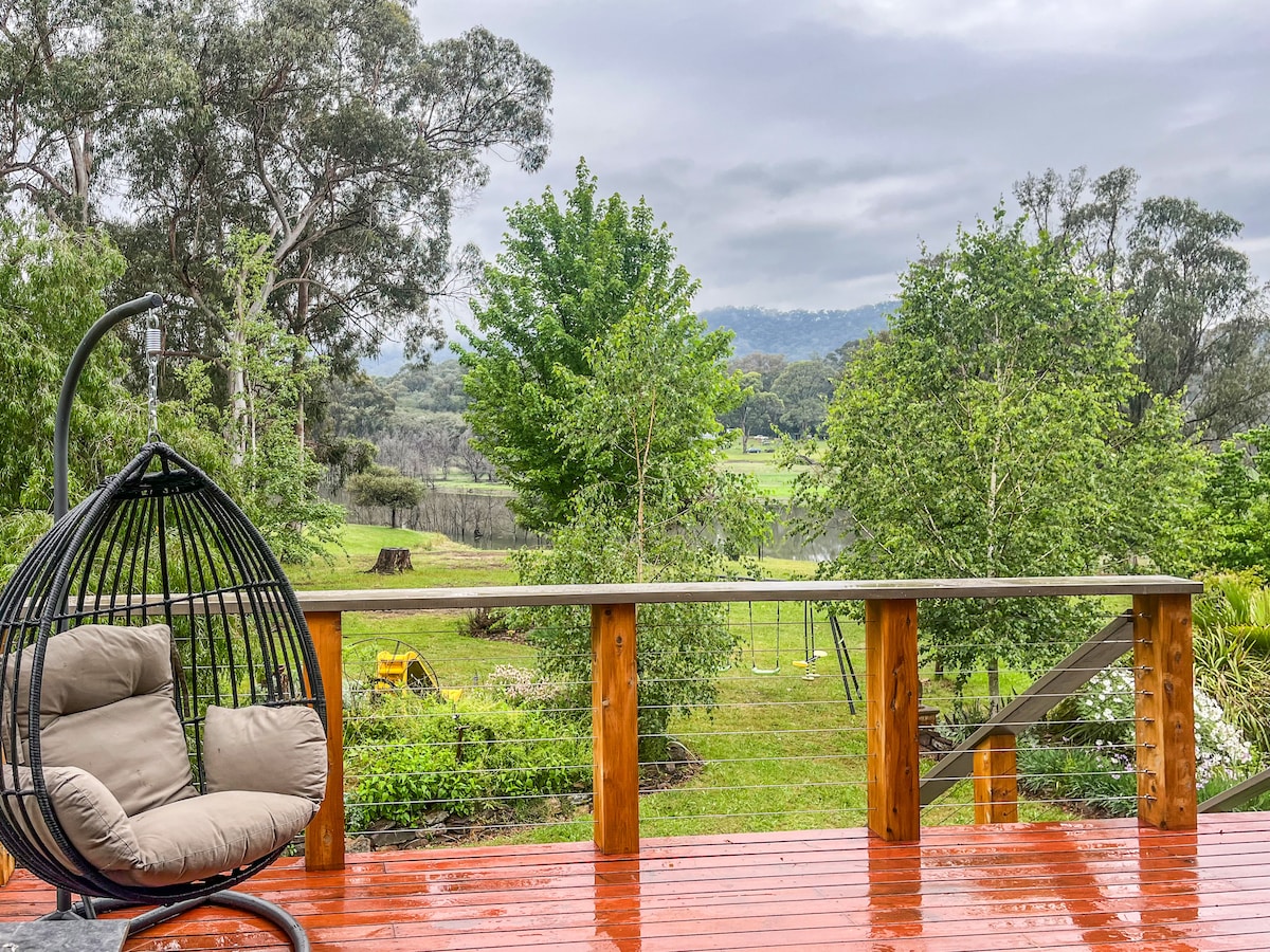 A hanging chair with plush cushions is positioned on a wooden deck, overlooking lush greenery and trees. The view reveals a serene landscape with gentle hills in the background, while the atmosphere is tranquil after a recent rain.