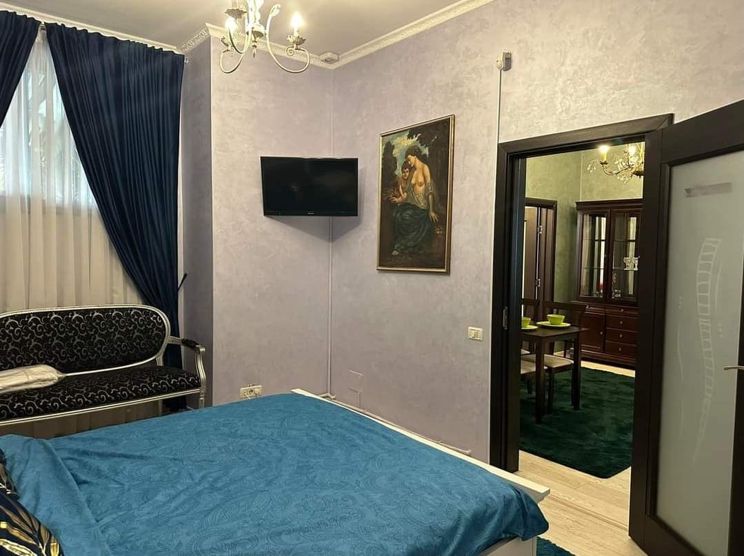 A cozy bedroom features a spacious bed covered in a blue blanket, with a television mounted on the wall. Elegant curtains frame the window, complementing the intricate artwork displayed nearby. The room is connected to an adjoining space furnished with a dark wood cabinet.