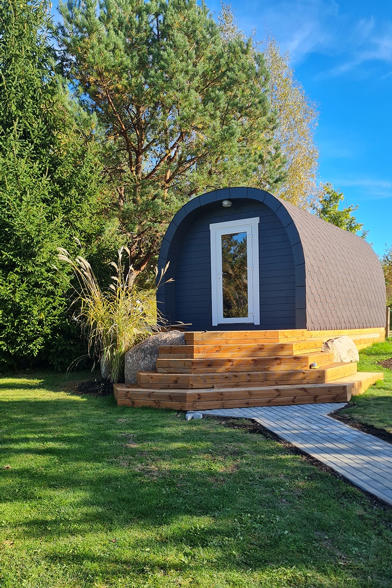 A compact, modern cabin is set against a backdrop of tall trees and greenery. Wooden steps lead to a glass door framed by large boulders. The cabin's curved roof and dark exterior create a unique silhouette under a clear blue sky.
