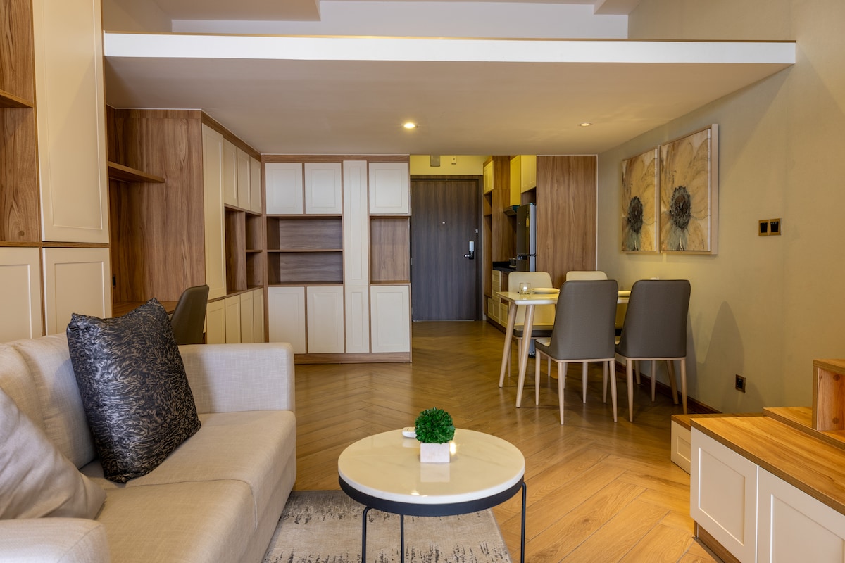 A cozy living area is presented with a light-colored sofa and a decorative cushion. A round coffee table sits in front, surrounded by warm wooden cabinets. In the background, a dining table with four chairs complements the modern kitchen, creating a functional living space.