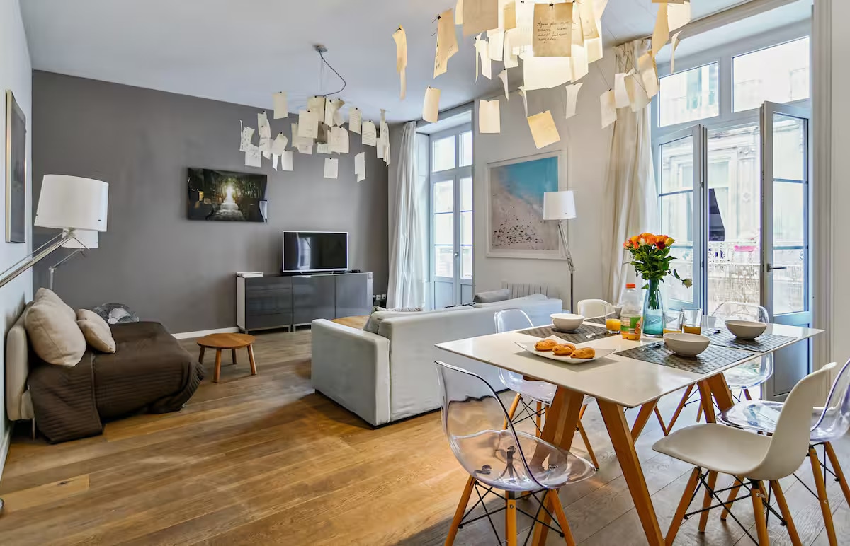 The spacious living area features large windows allowing ample natural light. A comfortable sofa and a stylish light fixture with hanging messages create a unique ambiance. A modern dining table with four white chairs is set with breakfast items, inviting social interaction.