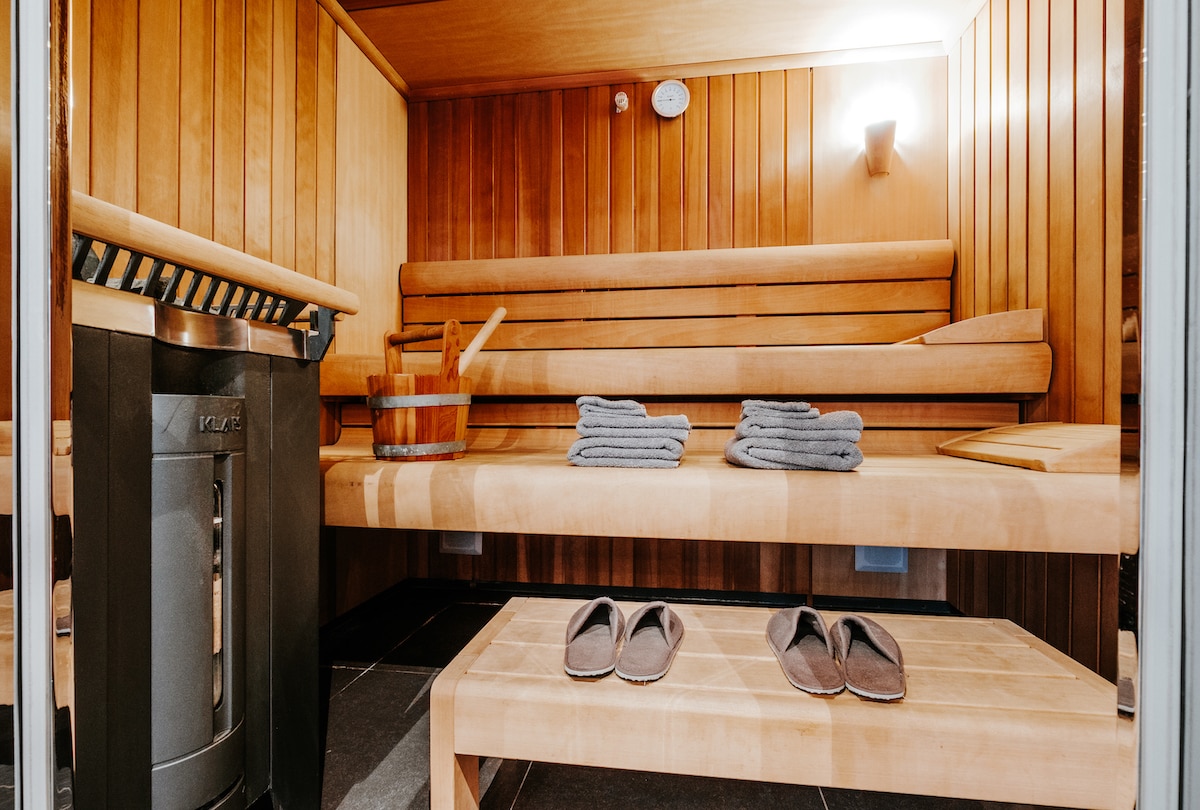 A warm wooden sauna is depicted, featuring two tiers of seating, a stone and wood heater, and a wooden bucket with a ladle. Soft towels are neatly arranged on the benches, while a pair of slippers reclines on a lower shelf, creating a calming atmosphere.