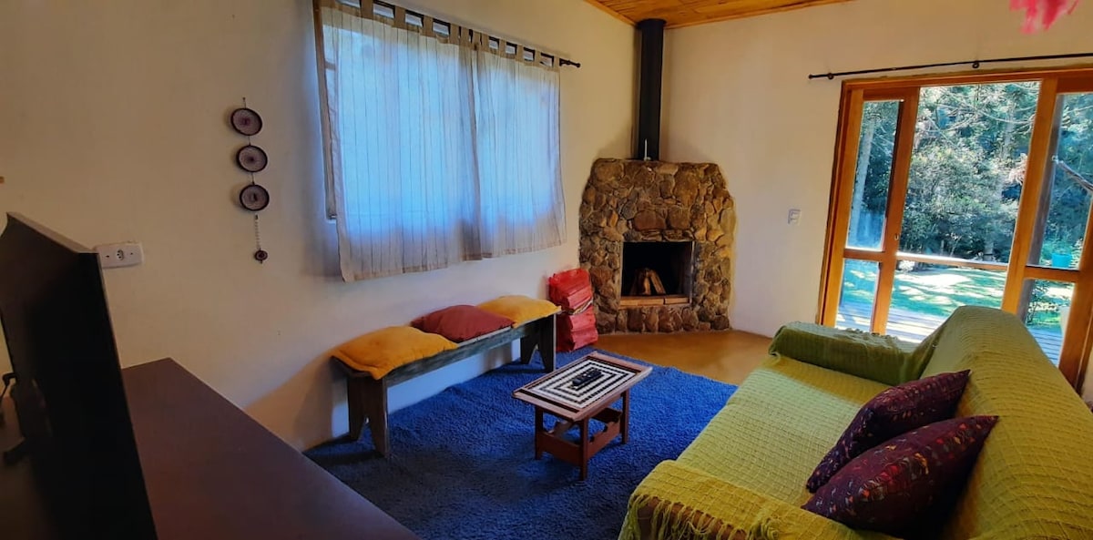 A rustic living area features a stone fireplace that adds warmth. A comfortable green sofa is positioned with vibrant cushions, accompanied by a wooden bench. Light filters gently through a large window, creating a bright and inviting atmosphere while providing a view of the green outdoors.