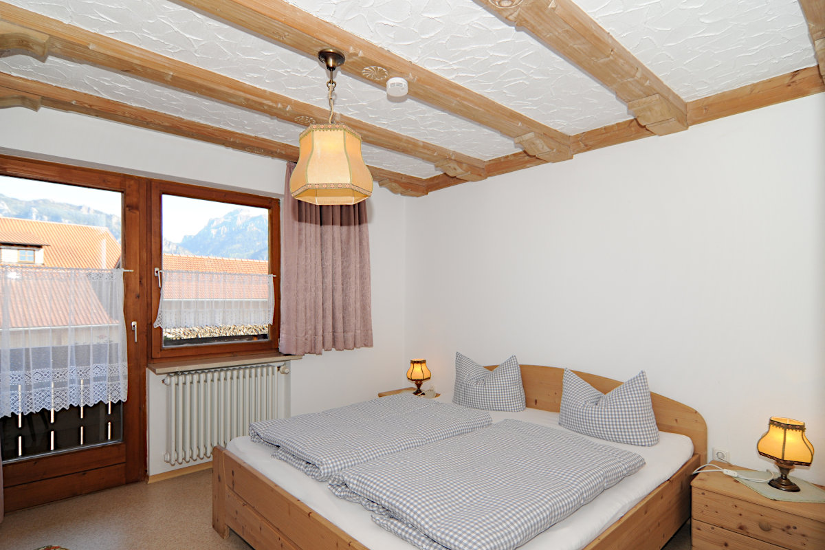 A well-lit bedroom features a spacious bed with light blue checkered bedding, complemented by wooden accents. Natural light enters through a window adorned with a sheer curtain, while soft lamps provide gentle illumination. Exposed wooden beams enhance the room's character.