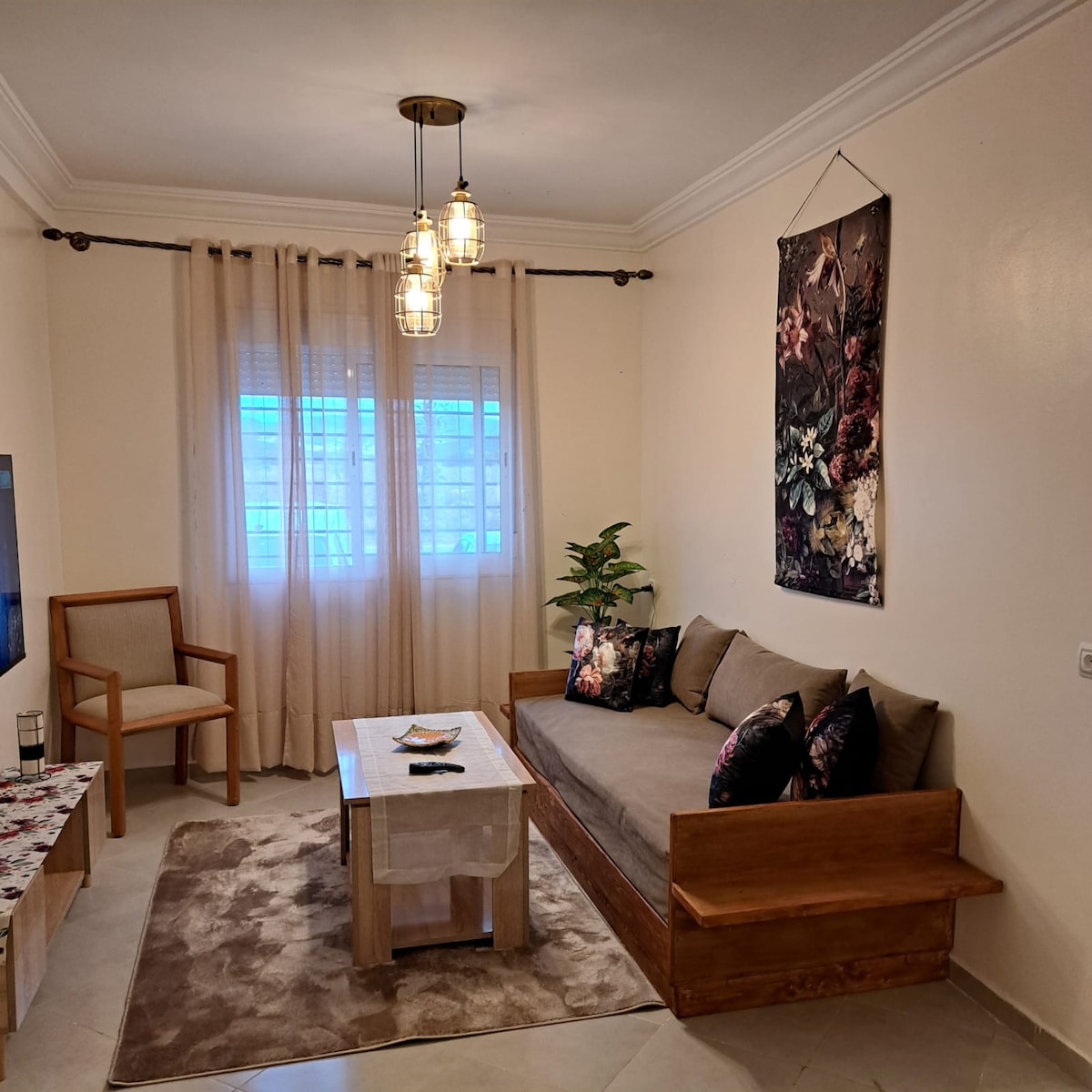 A living area features a convertible sofa adorned with decorative pillows, paired with a light wooden coffee table. Natural light filters through a window covered by sheer curtains, while a small plant adds a touch of greenery. A simple chair rests beside the sofa.