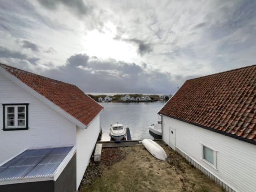 A serene waterfront view is presented, featuring two white houses with red-tiled roofs on either side. A calm body of water reflects the cloudy sky, with boats docked nearby. The surrounding area is tranquil and lined with greenery in the distance.