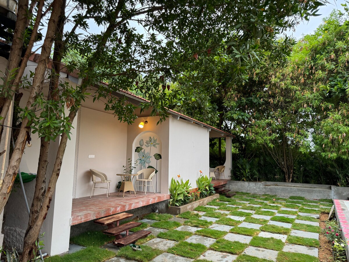 An outdoor seating area is revealed, framed by lush greenery. Two chairs and a small table are set on a brick patio, surrounded by a manicured lawn featuring square stone pavers. The building has light-colored walls adorned with a floral mural.