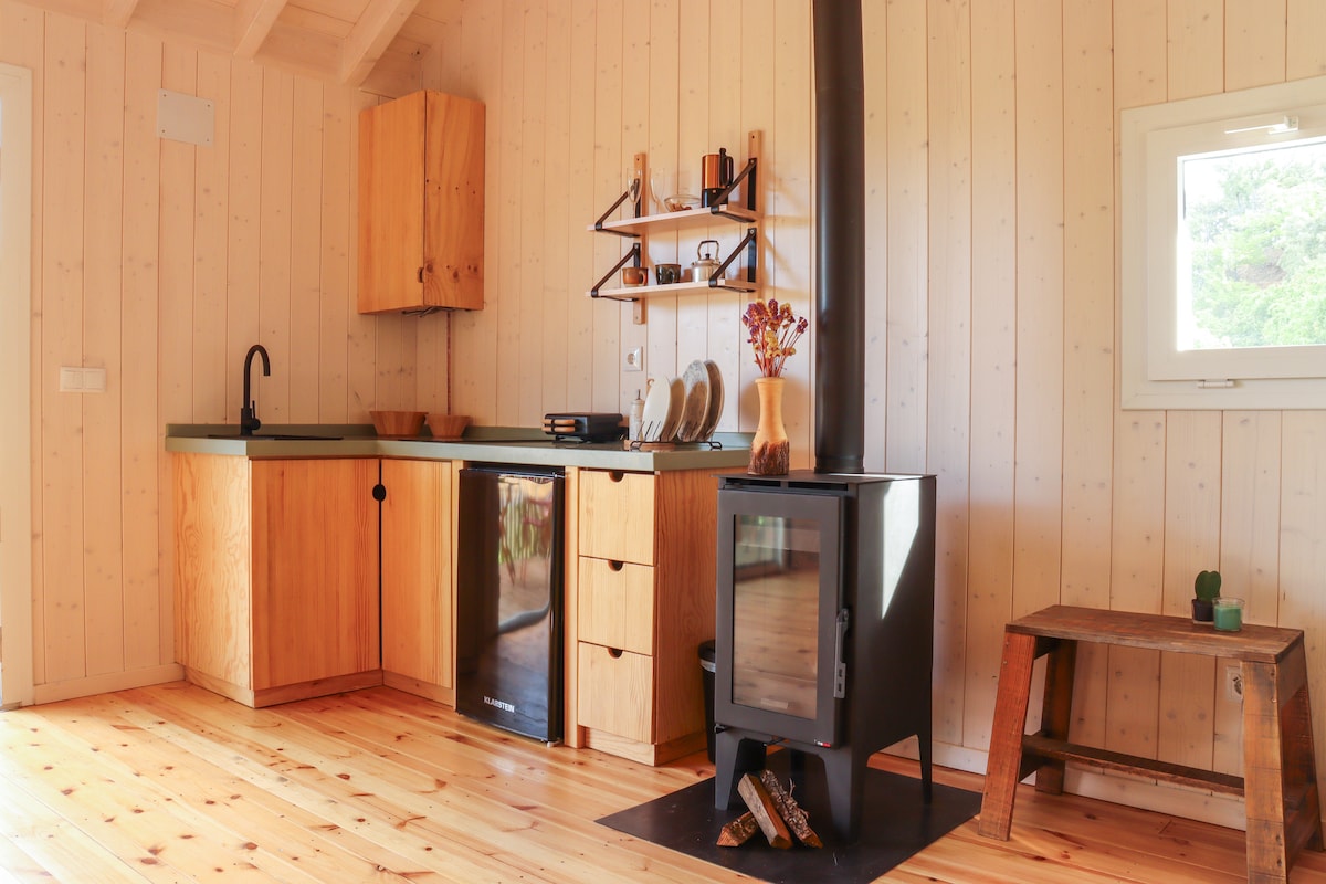 A functional kitchen area is highlighted by wooden cabinetry and a minimalist design. A black stove stands prominently on the wooden floor, accompanied by a small table and decorative items on the wall shelves. Natural light enters through a nearby window.