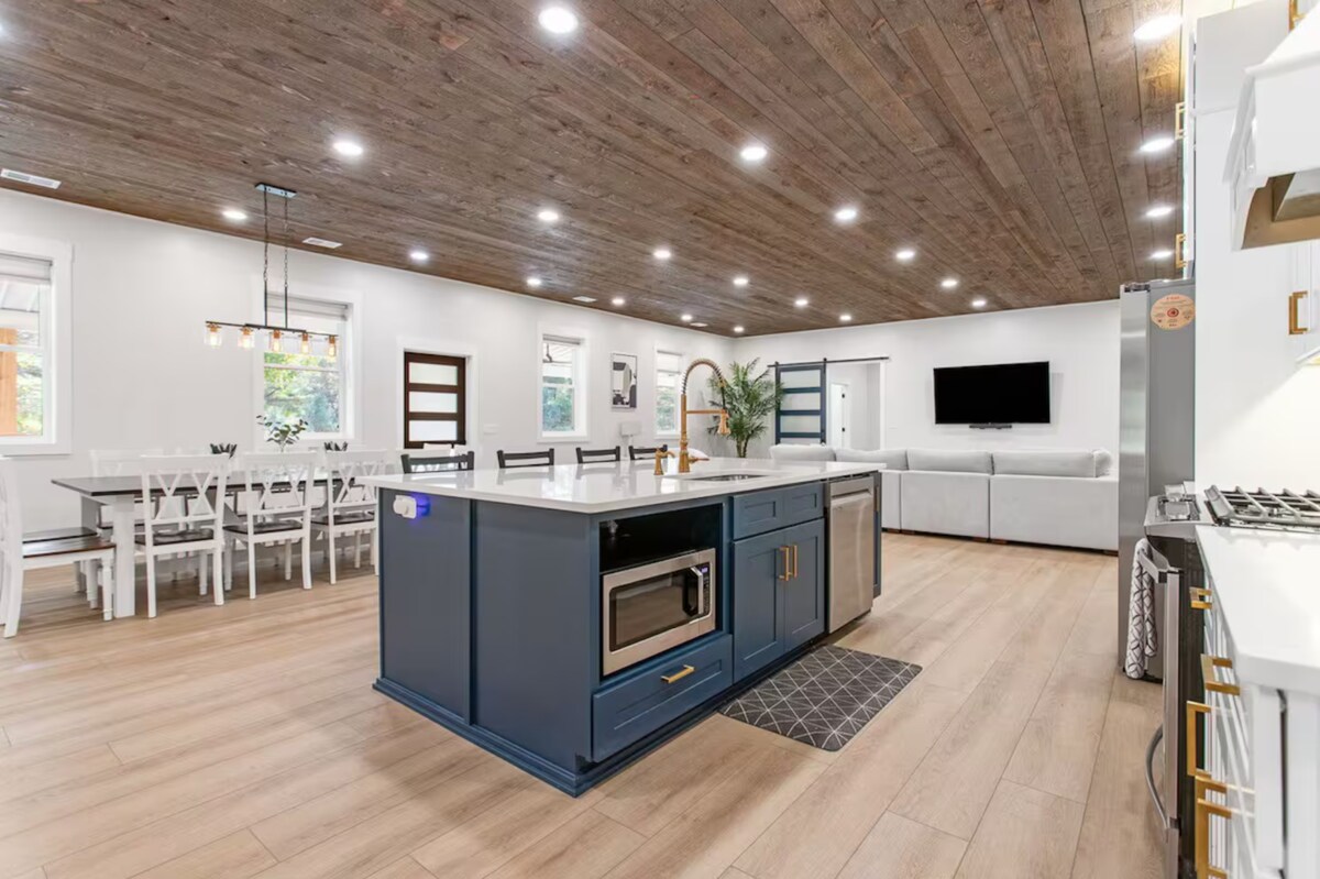 An open-concept kitchen and living area features a central island with a microwave and a dark blue finish. Light wood flooring contrasts with the wooden ceiling. A dining table for several diners is set beneath modern lighting, while a comfortable gray sectional sofa is positioned nearby.