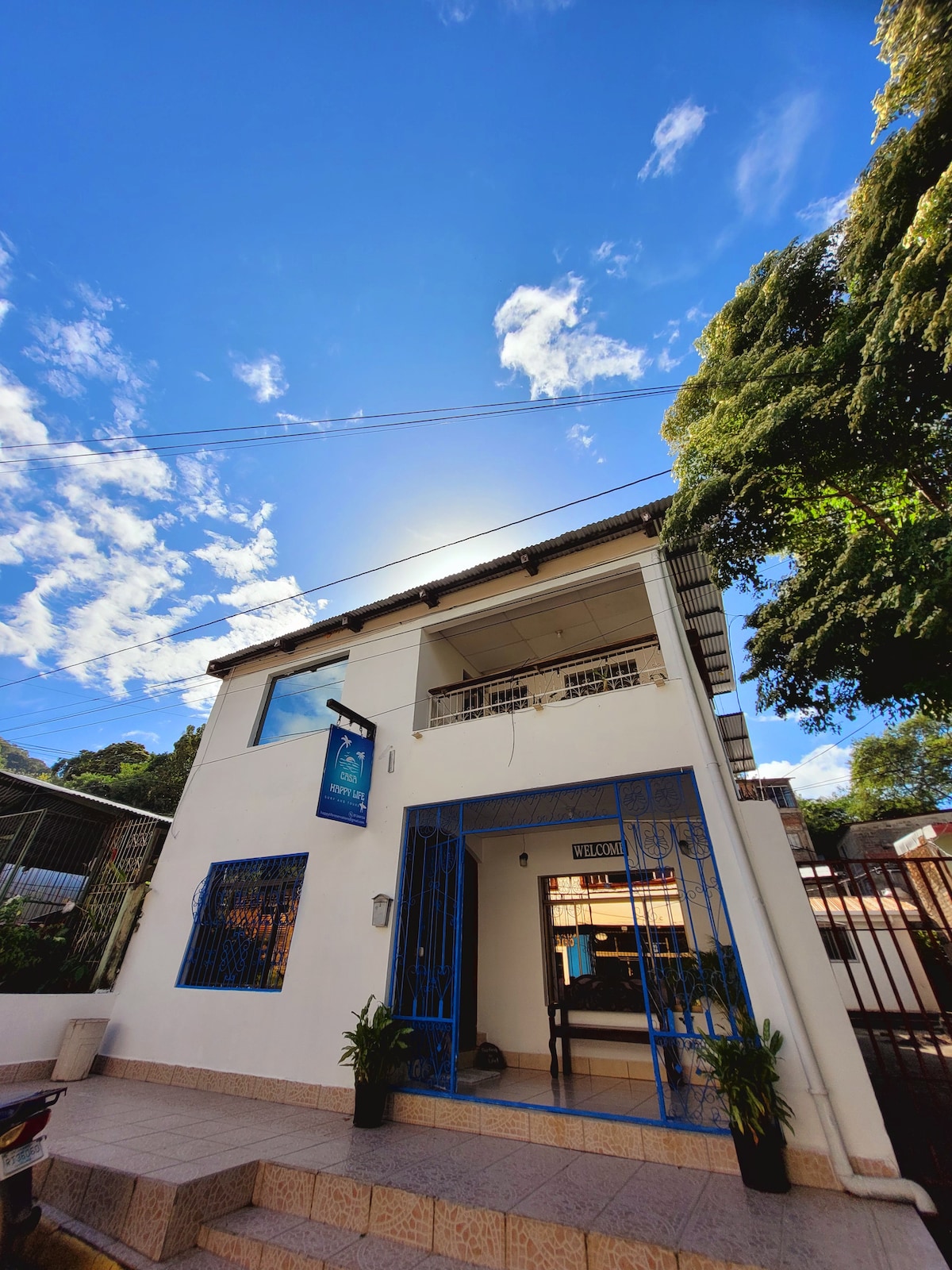 The entrance of a two-story building is shown under a clear blue sky. Bright steps lead to a welcoming doorway framed by vibrant blue wrought iron. Lush greenery is positioned alongside the entrance, contributing to the inviting setting.