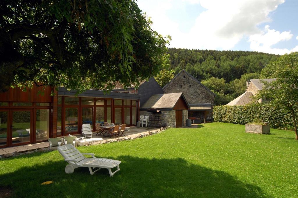 A spacious garden area is presented, featuring well-maintained grass and shaded seating. A sun lounger is positioned next to a patio with a wooden dining table and chairs, bordered by mature trees and a rustic stone building in the background.