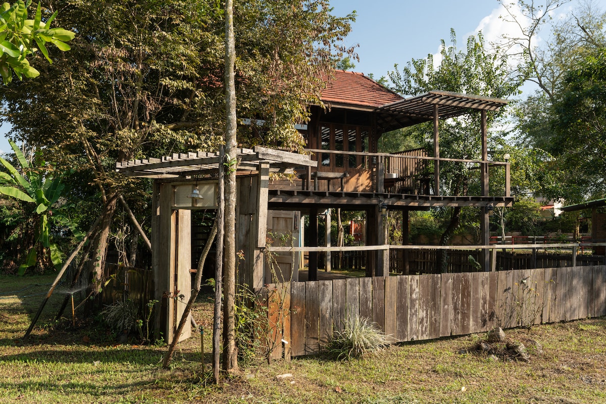 A multi-level countryside house is surrounded by lush greenery, showcasing wooden structures that blend harmoniously with the natural environment. A spacious balcony offers shaded outdoor seating, while a secure wooden fence creates a private garden area. Sunlight filters through the trees, enhancing the serene setting.