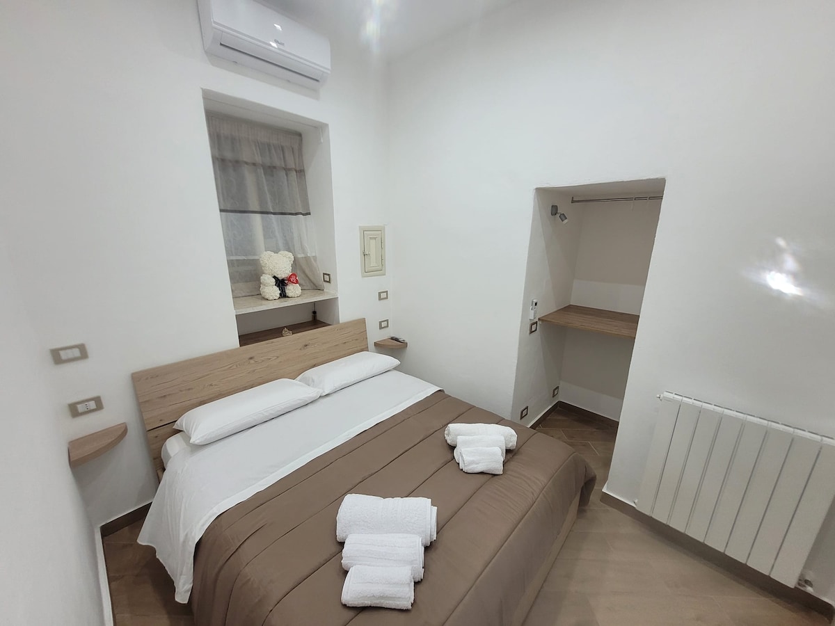 A well-appointed bedroom features a spacious bed with neutral linens and two neatly arranged towels. A soft teddy bear sits on the nightstand. A small desk area is visible, along with a window dressed in light curtains, allowing for natural light to filter in.