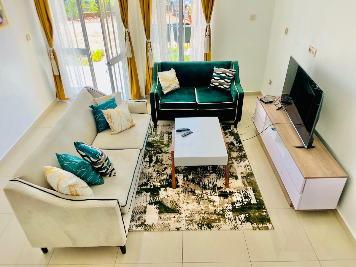 A living area features a light-colored sofa and a green velvet couch, each adorned with decorative cushions. A coffee table sits on a patterned rug, and a television is positioned on a low, modern media console. Natural light is allowed in through large windows, covered with elegant curtains.