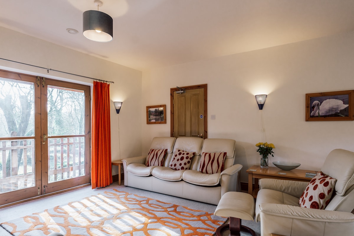 The living area features a comfortable seating arrangement, including a pair of armchairs and a sofa adorned with decorative cushions. Natural light floods in through double doors, leading to a balcony with forest views. Soft, warm lighting complements the cozy atmosphere.