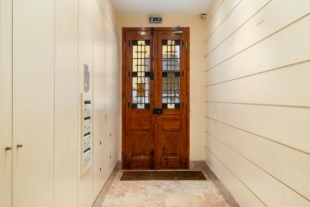 The entryway features a pair of large wooden doors with intricate glass panels framed by light-colored walls. To the left, a mail slot and a directory panel are visible, while a welcome mat is situated in front of the doors.
