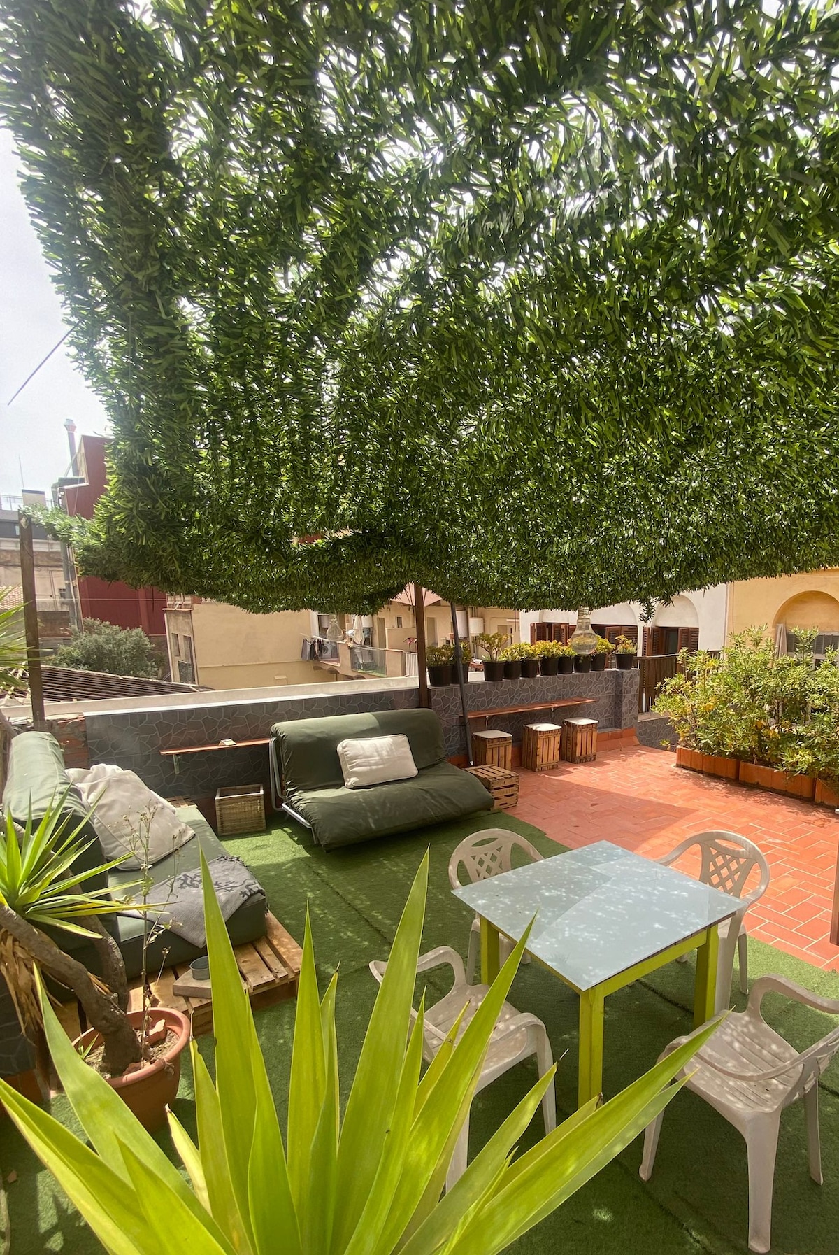 A private terrace is shaded by lush greenery, providing a serene outdoor setting. Comfortable seating areas feature soft cushions, while a small table and chairs create a space for dining. Potted plants add a touch of nature, enhancing the inviting atmosphere.