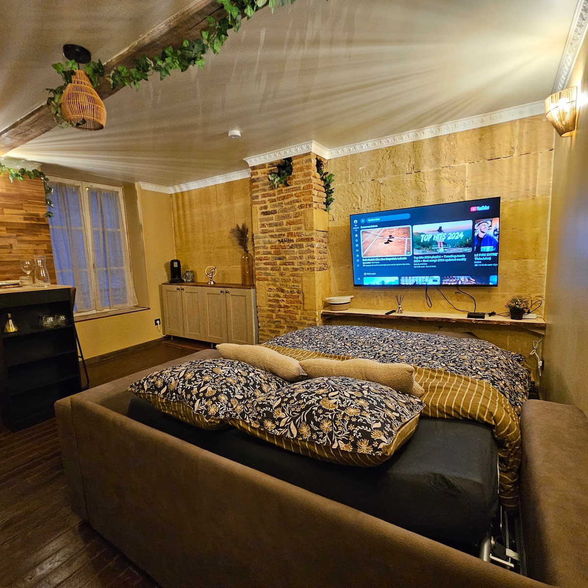 A studio space showcases a cozy sleeping area with a spacious bed adorned with patterned bedding. A large television is mounted on a brick wall, accompanied by a console below. Natural light enters through a window, enhancing the warm tones of the wooden accents.
