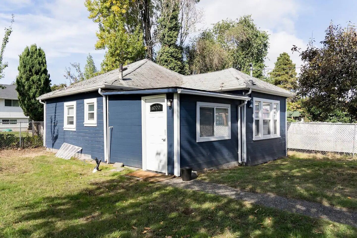 The exterior of a charming cottage is displayed, featuring a blue facade with multiple windows. A white door is visible, surrounded by grassy areas and a fenced backyard. Trees and greenery accompany the structure, contributing to a private outdoor space.