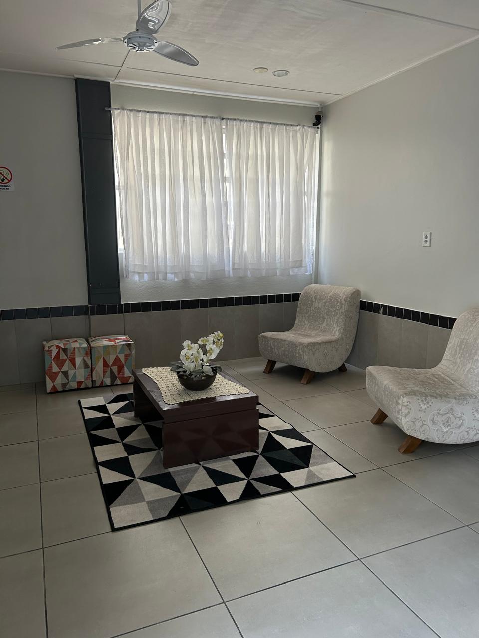 A welcoming living area features two light-colored chairs positioned around a low table. A patterned black and white rug lies on the tiled floor, with a bowl of flowers placed on the table. Natural light filters through sheer curtains.