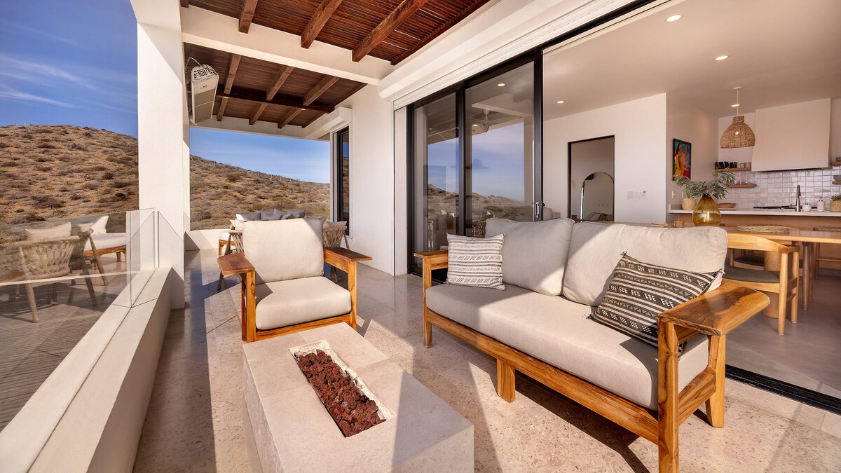An outdoor terrace showcases a comfortable seating area with wooden furniture. A small fire pit is centrally placed, surrounded by an expansive landscape of hills in the background. Large glass doors lead into a bright, open kitchen space.