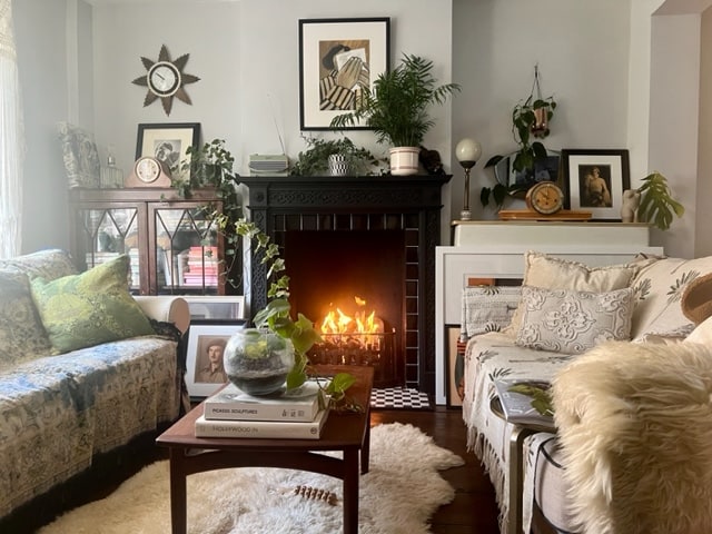 A cozy living room features a decorative fireplace with a gentle flame, surrounded by plants and framed art. Two comfortable sofas with patterned throws invite relaxation, while a small coffee table is adorned with books and a glass centerpiece, enhancing the room's tranquil ambiance.