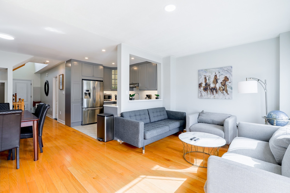 An open-concept living area showcases modern furnishings, featuring gray sofas and a round coffee table. Natural light floods in from large windows, illuminating the hardwood floors. The well-equipped kitchen, with gray cabinets and stainless-steel appliances, is visible in the background, creating a seamless flow between spaces.
