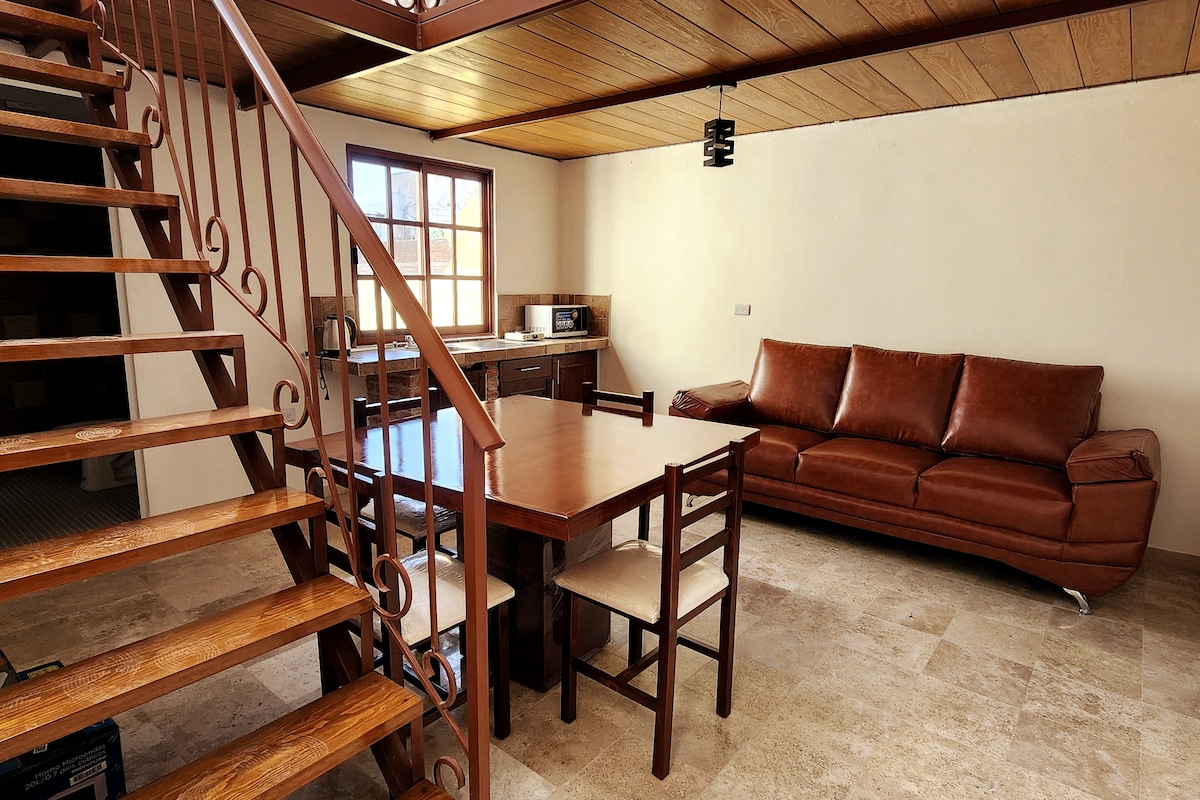 An open living space features a wooden dining table surrounded by four chairs and a spacious brown leather sofa. A staircase with ornate railings leads to an upper level. Natural light fills the room through a large window, highlighting the beige walls and tiled flooring.