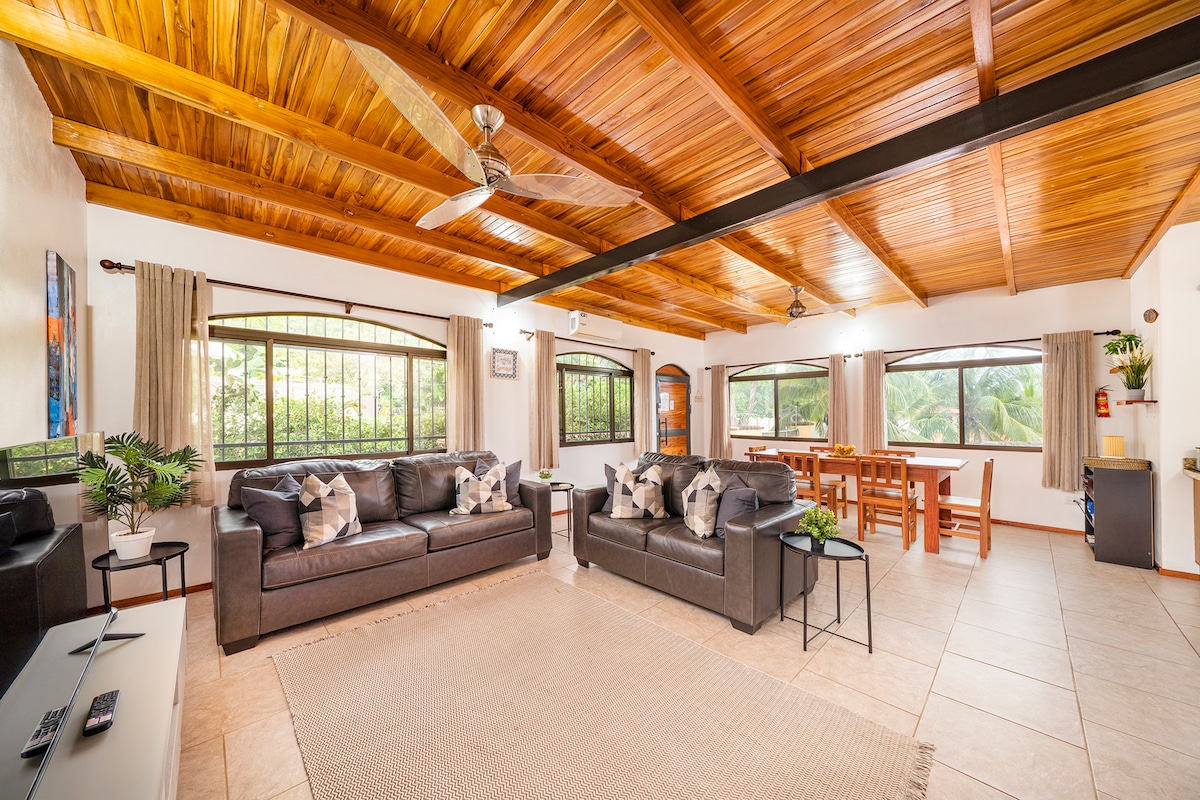The spacious main living area features dark leather sofas and a light-colored rug, providing a comfortable gathering space. A wooden ceiling and large windows allow natural light to brighten the room while offering views of the surrounding greenery. A dining table is positioned in the background.