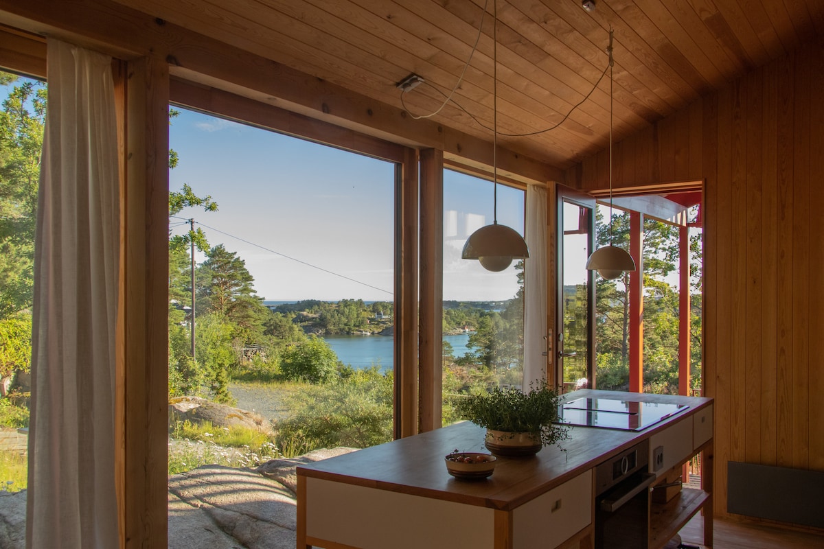 A spacious kitchen area is highlighted by large windows that offer stunning views of the surrounding landscape and water. Natural wood paneling adds warmth, while two pendant lights hang above a central island. Light, sheer curtains softly frame the windows.