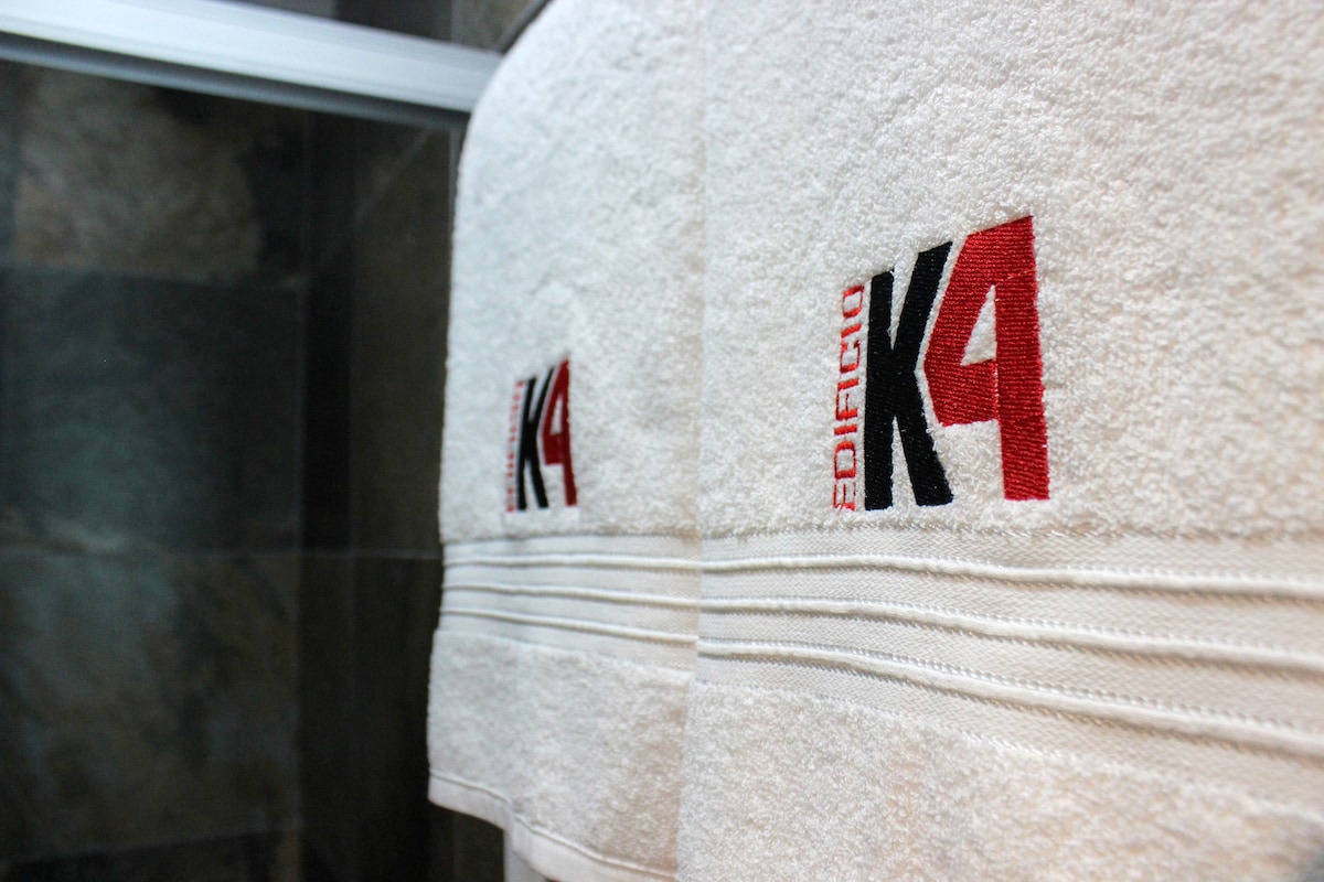 Soft, white towels are displayed neatly, featuring an embroidered logo in red and black. The towels are hung on a bar, surrounded by dark stone walls, contributing to a clean and sophisticated bathroom environment.