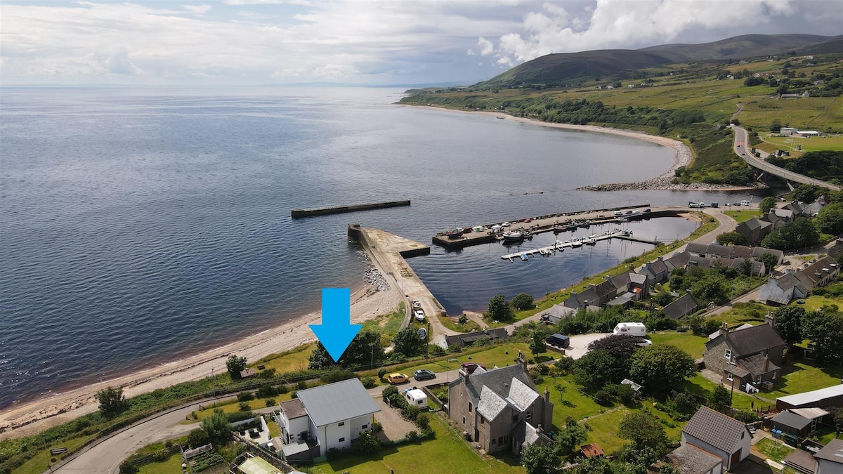 An aerial view captures the tranquil coastal setting, highlighting the nearby beach and marina. The shoreline is visible, along with several structures dotting the landscape and the expansive water of the Moray Firth stretching into the distance under a partly cloudy sky.