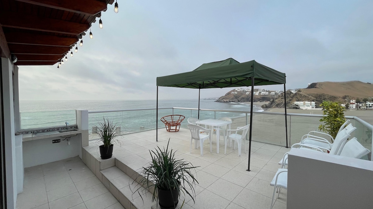 A spacious terrace overlooks the ocean, featuring a green canopy shade structure and a dining table with white chairs. Potted plants are positioned along the edge, offering a touch of greenery against the neutral tiles and the scenic beach view.