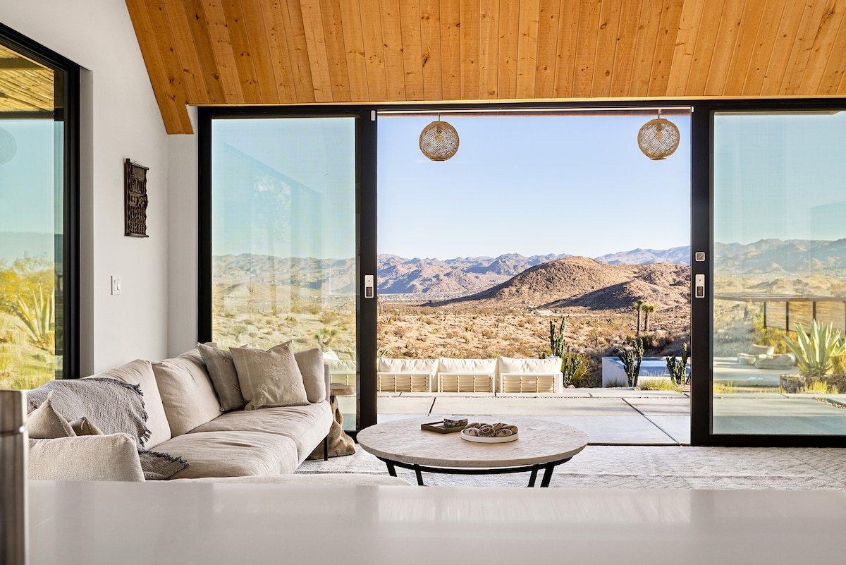 A modern living area is highlighted by large glass doors that open to breathtaking desert views. A light-colored sofa is positioned beside a natural wood coffee table, inviting relaxation. Wood-paneled ceilings add warmth, while the surrounding landscape enhances the spacious feel.