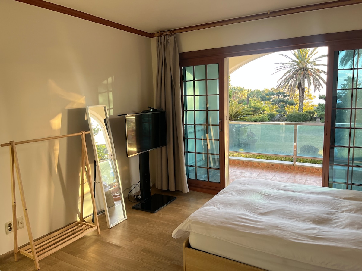 A sleeping area features a spacious bed, illuminated by natural light from sliding glass doors that open to a scenic view. A television stands on a stand opposite the bed, while a mirror and a wooden clothing rack are positioned nearby.
