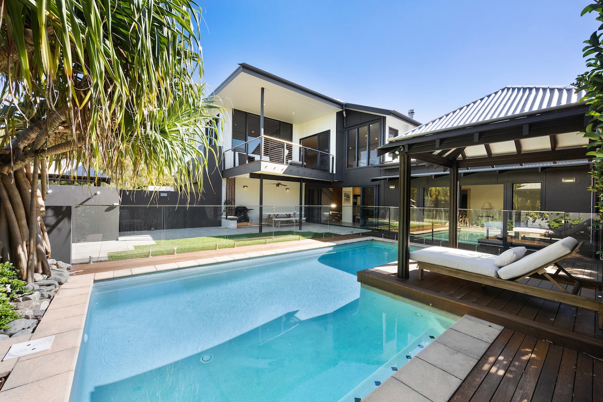 A modern beach house is showcased, featuring a sparkling inground pool bordered by grass and a wooden deck. Lush palm trees provide shade, while the expansive balcony above overlooks the pool area. The structure is designed with clean lines and large glass panels, enhancing natural light.