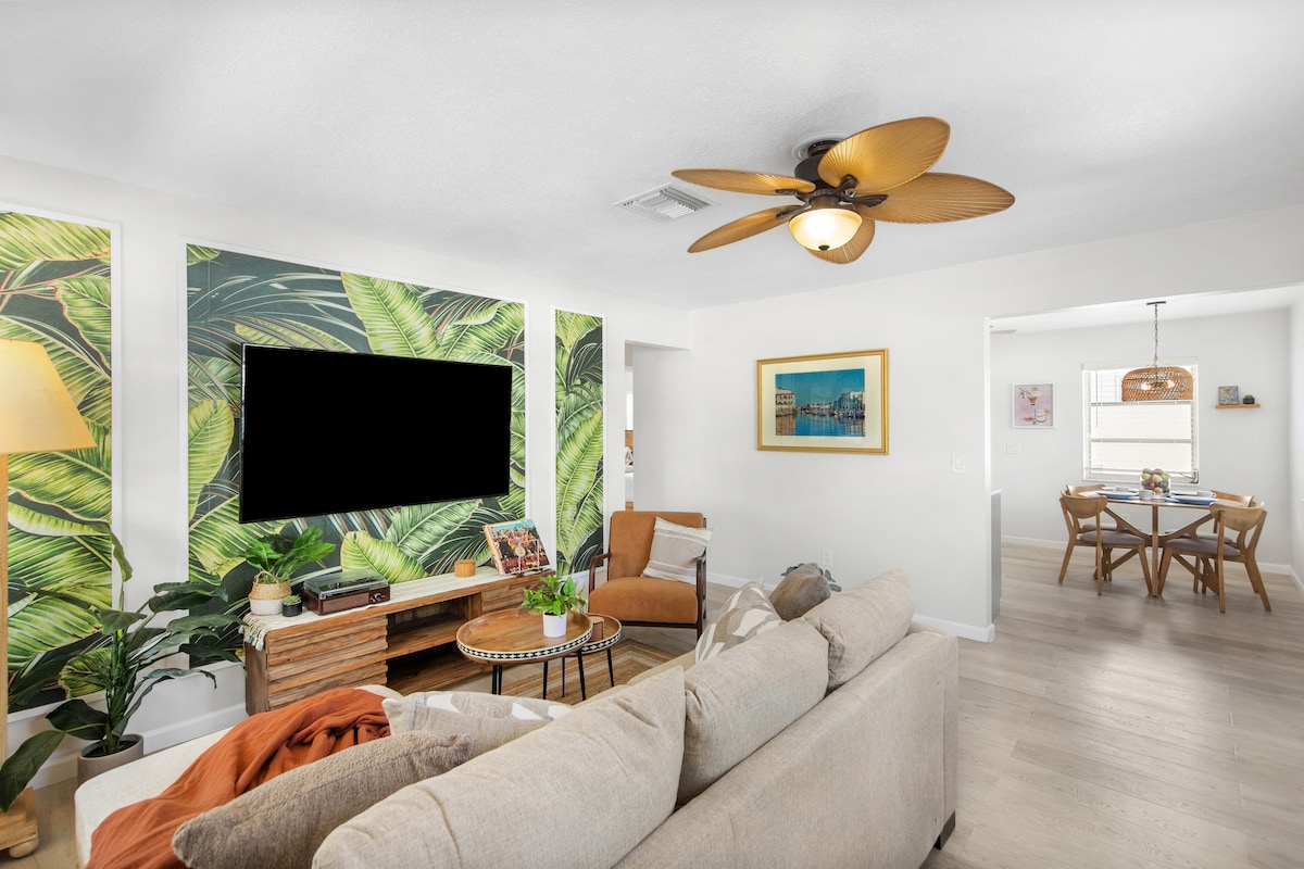 A cozy living room features a couch and armchair, with a coffee table centered in front. One wall is adorned with tropical leaf wallpaper, while a flat-screen TV is mounted above a wooden media console. A dining area with a round table is visible in the background.