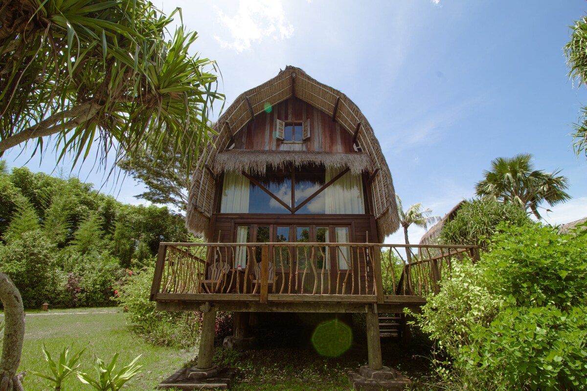 A charming retreat is showcased, featuring a thatched roof and wooden accents. Large glass windows invite natural light, while a wooden veranda extends from the front. Lush greenery surrounds the structure, with tropical plants framing the pathway leading to the entrance.