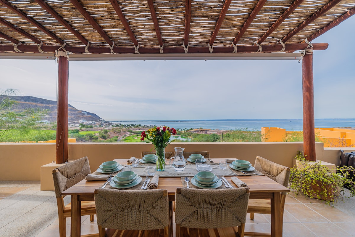 An outdoor dining area features a wooden table set for six, adorned with plates and glassware. Overhead, a thatched roof provides shade, while stunning ocean views extend into the distance. Potted plants and a gentle breeze enhance the serene atmosphere.