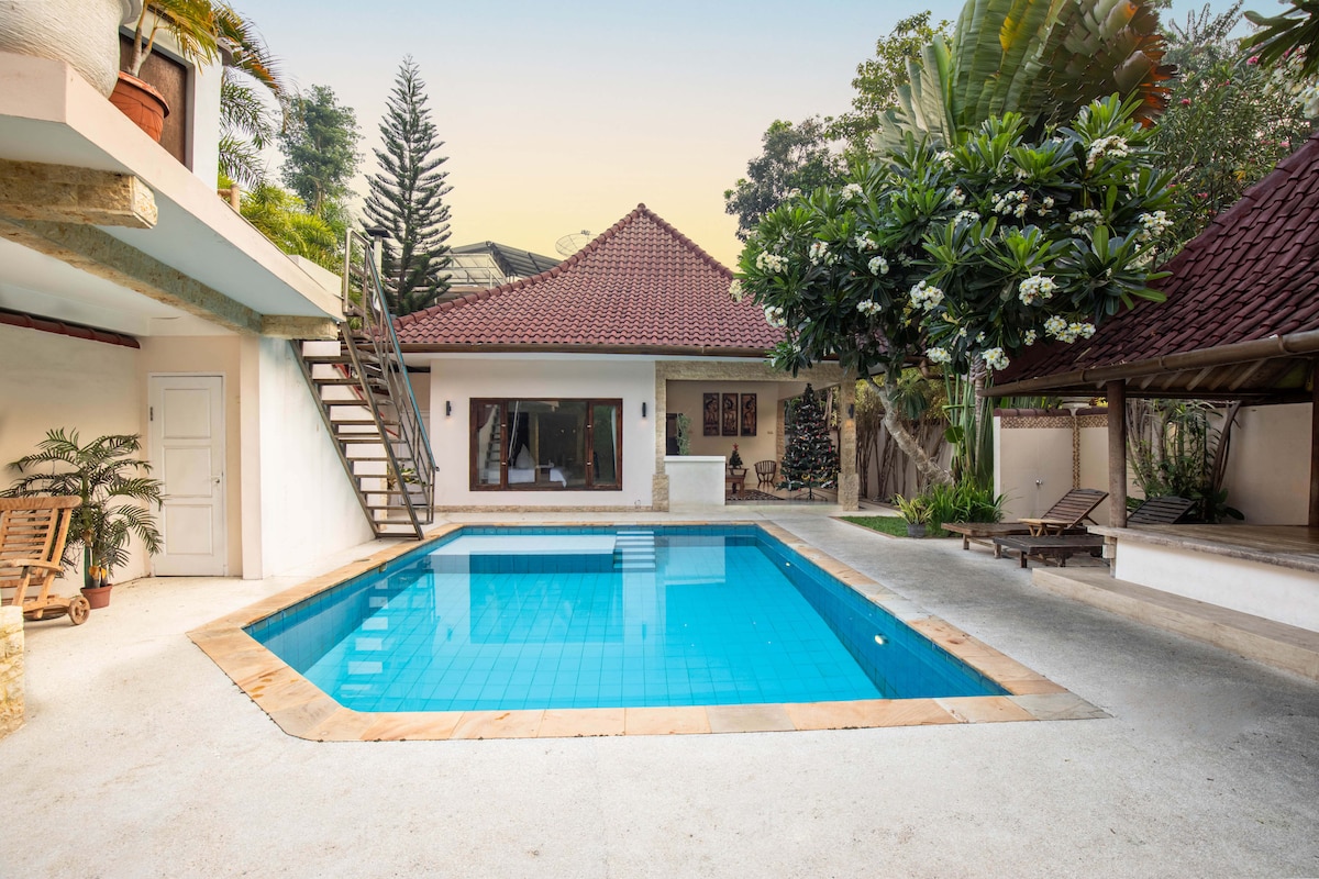 A private pool is centrally featured within a spacious outdoor area, surrounded by lush greenery and tropical plants. The villa's exterior is framed by a traditional roof, with an inviting seating area visible to the side. A staircase leads to an upper level.