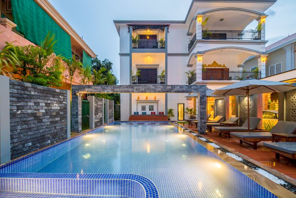 A modern villa is featured with a large, inviting swimming pool that reflects the surrounding plants and structures. Comfortable loungers are positioned nearby, offering a relaxing space. The building showcases a blend of architectural styles with balconies and decorative elements illuminated in the early evening light.
