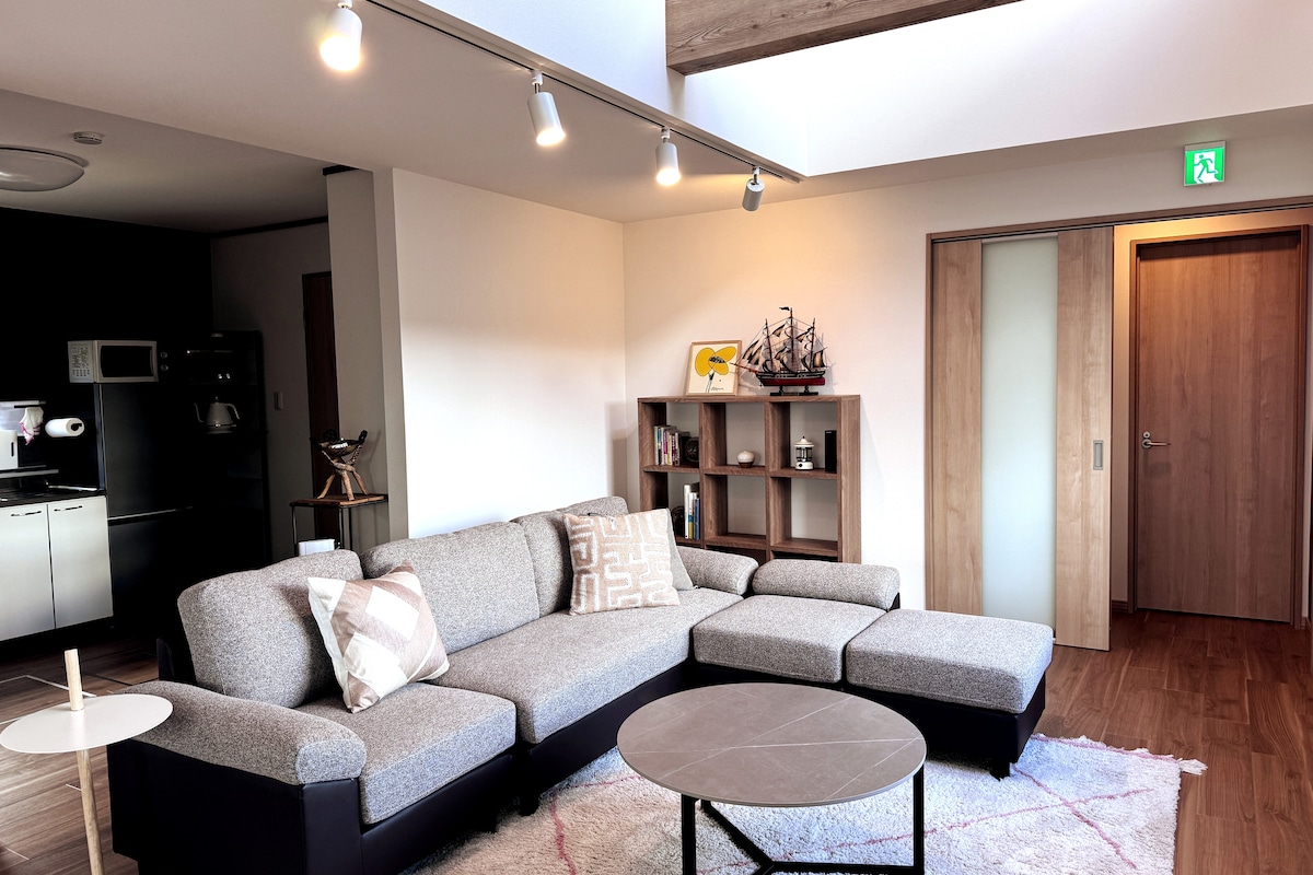 A spacious living area features a comfortable gray sectional sofa and a round coffee table. Natural light is allowed in through windows, and a decorative shelf holds small sculptures and a model ship. The ceiling showcases track lighting, illuminating the space softly.