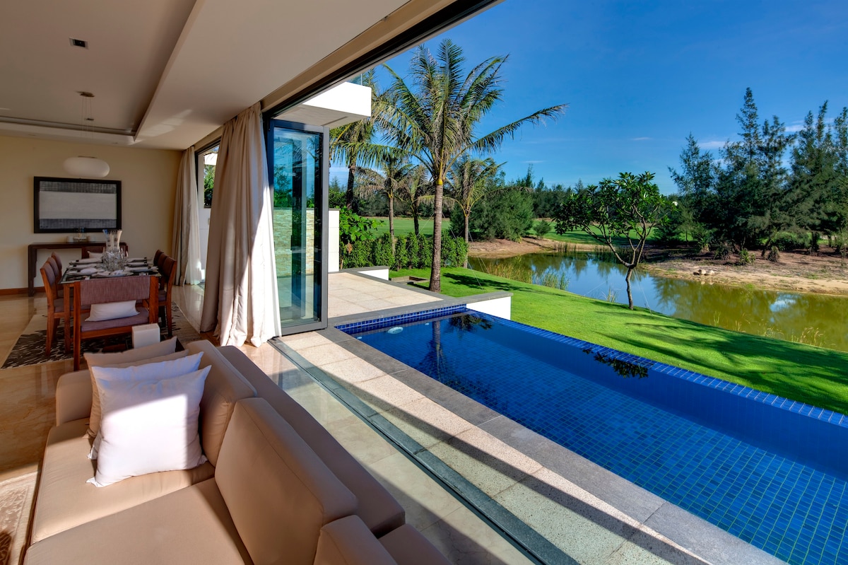 A spacious living area is illuminated by natural light from large glass doors. An inviting outdoor swimming pool extends from the lounge, positioned alongside a well-maintained garden and water feature, surrounded by lush greenery and palm trees.