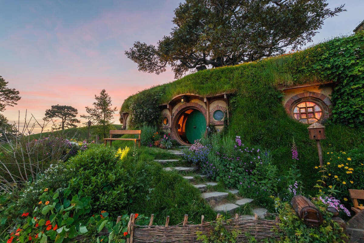 A charming cottage entryway is framed by lush green grass and vibrant flowers. Stone steps lead to a round door, surrounded by small, circular windows. A wooden bench is positioned beside the path, inviting visitors to enjoy the peaceful garden setting.