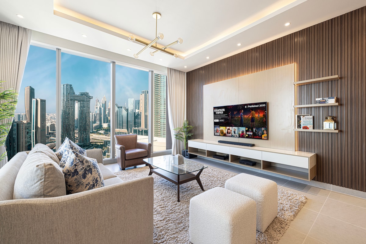 A bright living area is revealed, featuring large windows that offer panoramic views of the city skyline. A soft rug covers the floor, complemented by a cozy couch and modern armchairs. A sleek entertainment unit houses a television, and decorative elements add a touch of style.