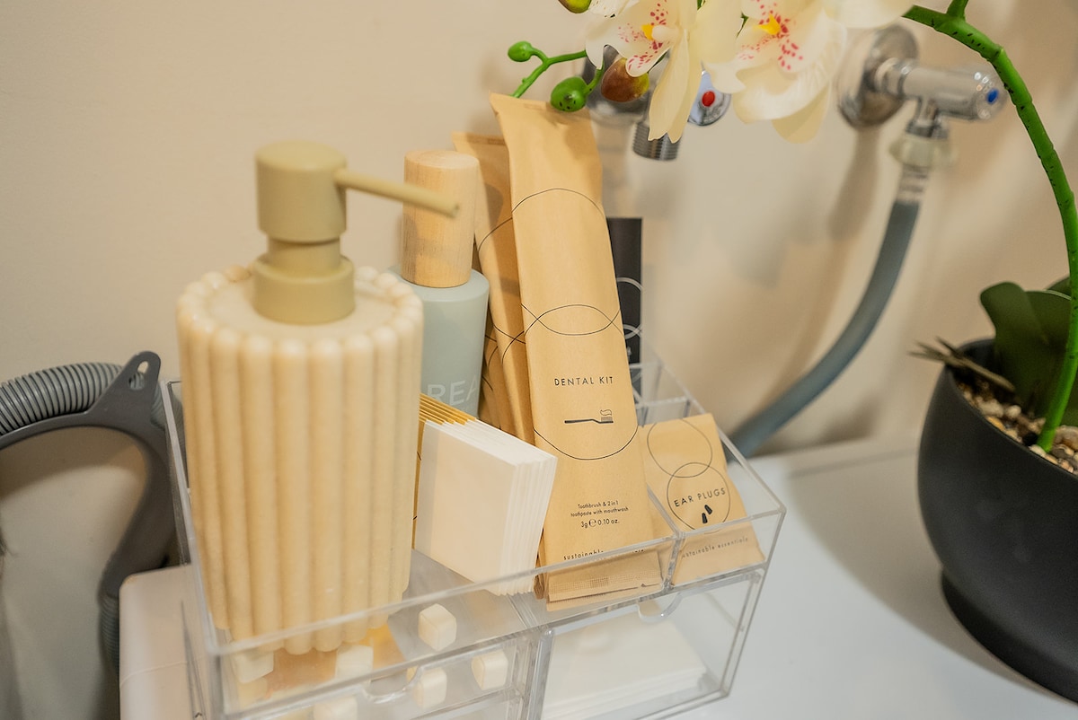 A well-organized bathroom counter displays a collection of personal care items. A textured pump dispenser is placed beside a dental kit, complemented by neatly packaged toiletries and cotton swabs. An elegant orchid adds a touch of greenery to the arrangement.